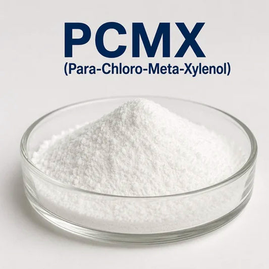 A small clear glass dish filled with white powder, with large blue text above reading 'PCMX (Para-Chloro-Meta-Xylenol)' on a light gray - Hbackground.