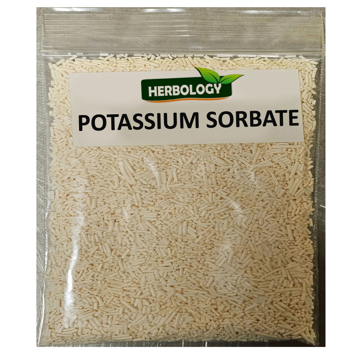 A clear plastic resealable bag filled with light beige potassium sorbate granules, featuring a white label with the green and orange 'Herbology' brand logo and the text 'POTASSIUM SORBATE'.